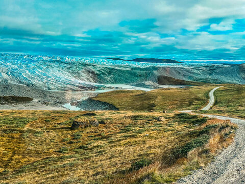 View of the vast arctic landscape featuring a massive glacier, rugged tundra terrain, and a winding dirt road under a dramatic cloudy sky in Greenland.