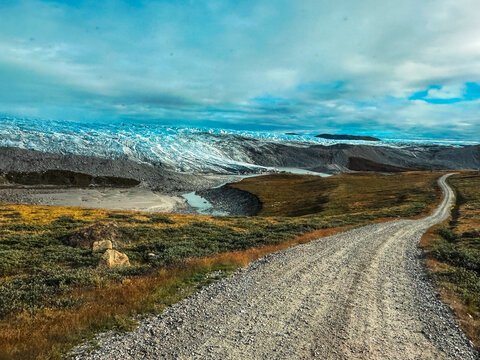 View of a gravel road winding through the vast tundra landscape towards a massive glacier under a dramatic cloudy sky in Greenland.