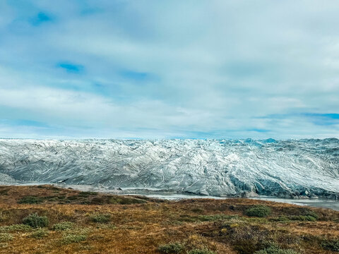 View of the expansive icy surface of a glacier with rugged crevasses and textures, set against a backdrop of tundra and a cloudy sky in Greenland.