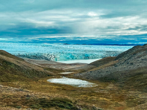 View of a massive blue glacier flowing into a glacial lake surrounded by rugged rocky hills and tundra under a cloudy sky in Greenland.
