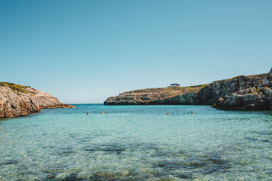 View of Cala Binidali with turquoise water, rocky cliffs, and people swimming in the cove in Mao, Illes Balears, Spain.