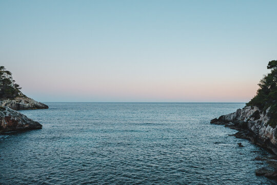 View of the calm waters of Cala Galdana bay framed by rocky cliffs under a clear sky at dusk, Cala Galdana, Illes Balears, Spain.
