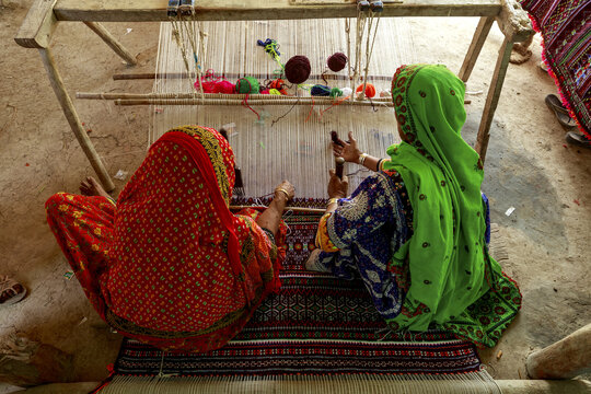 View of two women wearing traditional colorful clothing working on a traditional loom to create a farasi in Badin, Sindh, Pakistan.