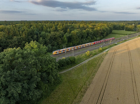 Aerial view of a passenger train traveling on tracks through a lush green forest and a golden wheat field in Germany.