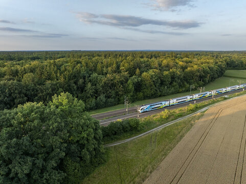 Aerial view of a modern passenger train traveling on tracks through a lush forest and golden wheat fields in the rural countryside of Germany.