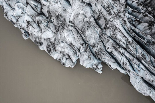 Aerial view of a glacier with deep crevasses and dark volcanic ash deposits meeting a glacial river in Iceland.