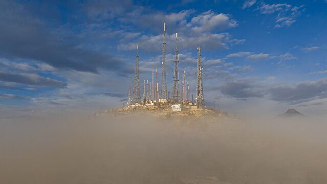 Aerial view of Akyokus hill featuring multiple telecommunication towers rising above a thick layer of morning fog under a blue sky in Meram, Konya, Turkiye.