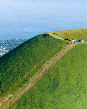Aerial view of Mt. Omuro, a lush green volcanic cone with a chairlift line ascending the slope and the blue sea in the background in Ito, Shizuoka, Japan.