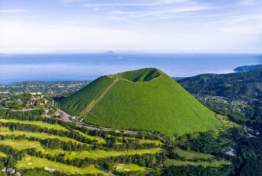 Aerial view of Mt. Omuro, a prominent volcanic cinder cone with lush green slopes and a crater, surrounded by a golf course and the ocean in Ito, Shizuoka, Japan.