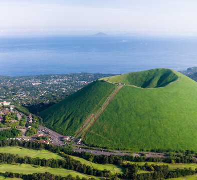 Aerial view of Mt. Omuro, a dormant volcanic cone covered in lush green grass, rising above the coastal town and the blue waters of the Pacific Ocean in Ito, Shizuoka, Japan.