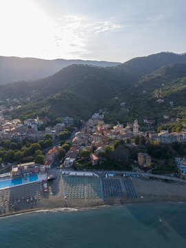 Aerial view of a coastal town with colorful houses, a church bell tower, and a sandy beach with rows of umbrellas nestled against lush green hills in Italy.