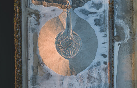 Aerial view of a circular sand stockpile with striking radial excavation patterns, featuring contrasting warm and cool tones in an industrial landscape in France.