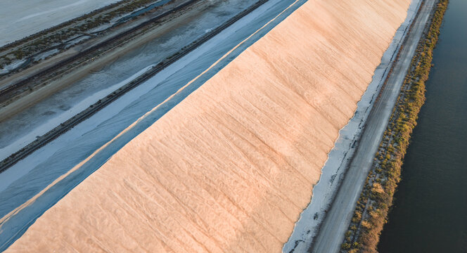 Aerial view of giant white salt mounds in the Camargue salt marshes, featuring geometric patterns created by warm evening light and cool blue shadows, France.