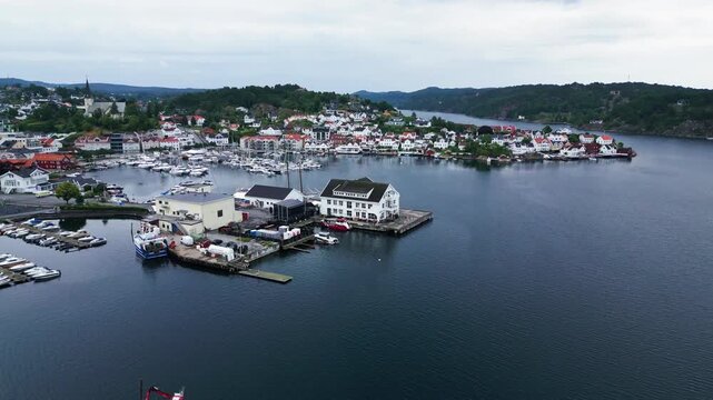 Aerial view of the coastal town of Grimstad with its harbor filled with boats, white wooden houses, and surrounding hills in Grimstad, Agder, Norway.