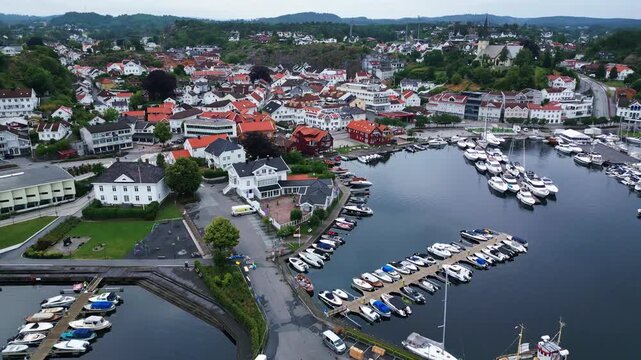 Aerial view of the coastal town of Grimstad with its marina filled with boats, white wooden houses, and lush green surroundings in Grimstad, Agder, Norway.