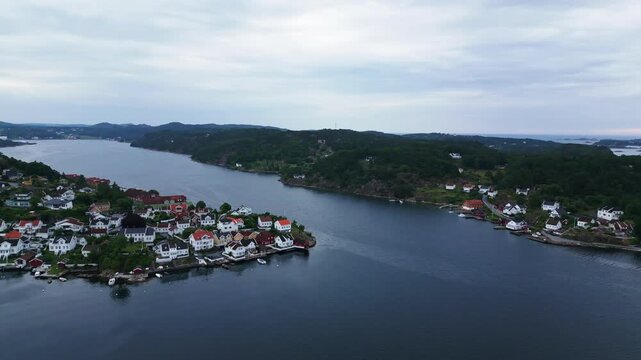Aerial view of the coastal town of Grimstad with traditional white houses along the waterfront, surrounded by lush green hills and calm blue water in Grimstad, Agder, Norway.