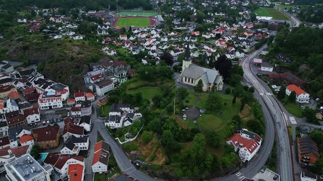 Aerial view of the historic Grimstad Church surrounded by residential houses, lush green trees, and winding roads in the coastal town of Grimstad, Agder, Norway.