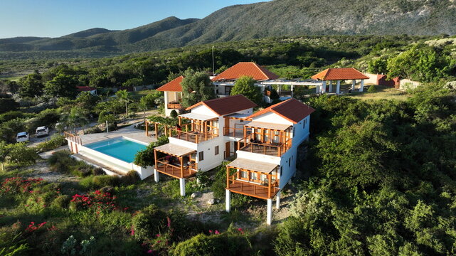 Aerial view of Altos de La Caobita Eco-hotel with white buildings, red roofs, wooden balconies, and a swimming pool set in lush green vegetation in Barrera, Azua, Dominican Republic.