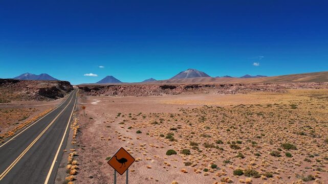 Aerial view of a paved road winding through the arid Atacama Desert landscape with a rhea warning sign and distant volcanic mountains in Antofagasta, Antofagasta, Chile.