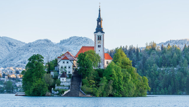 View of the Pilgrimage Church of the Assumption of Maria on the island in Lake Bled, surrounded by green trees and snow-capped mountains in the background, Bled, Radovljica, Slovenia.