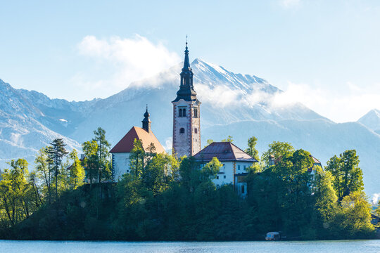 View of Lake Bled island with the Pilgrimage Church of the Assumption of Maria, surrounded by trees, with snow-capped Julian Alps in the background, Bled, Radovljica, Slovenia.