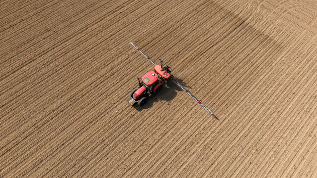 Aerial view of a red tractor with a sprayer boom working in a large cultivated field with brown soil furrows in Mediglia, Lombardy, Italy.