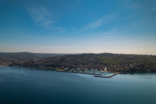 Aerial view of Muggia marina with boats docked in the harbor, surrounded by the coastal town and lush green hills under a clear blue sky in Trieste, Friuli-Venezia Giulia, Italy.