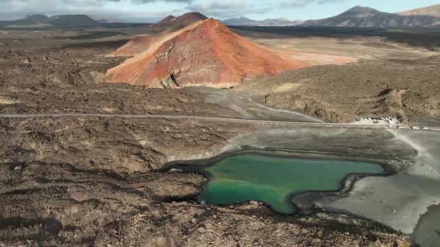 Aerial view of the green Charco de los Clicos lagoon surrounded by black lava fields and a red volcanic cone in Lanzarote, Spain.
