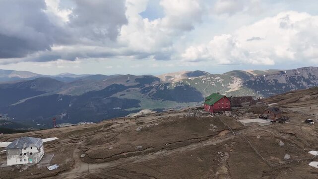Aerial view of the rugged Bucegi Mountains landscape featuring a mountain hut and cable car infrastructure under a cloudy sky in Sinaia, Prahova, Romania.