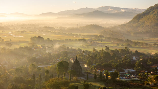 Aerial view of Sojiwan Temple surrounded by lush greenery, misty fields, and distant mountains during golden hour in Kecamatan Prambanan, Jawa Tengah, Indonesia.