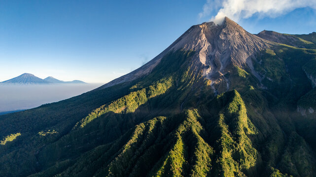 Aerial view of Mount Merapi with smoke rising from the crater, lush green slopes, and distant mountains under a clear blue sky, Kecamatan Cangkringan, Daerah Istimewa Yogya, Indonesia.