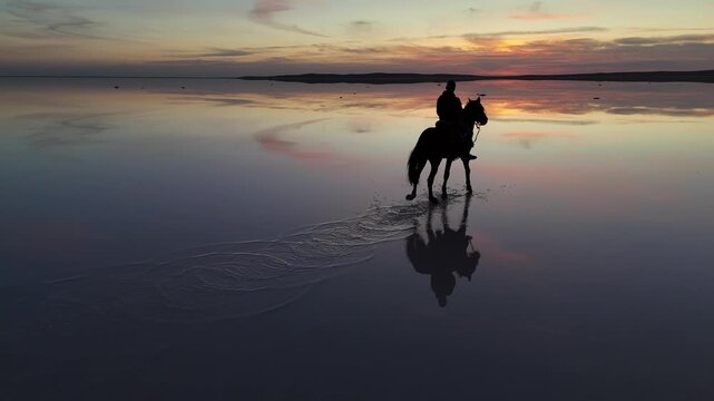 Aerial view of a person riding a horse through the shallow waters of Lake Tuz at sunset with vibrant sky reflections in Kulu, Konya, Turkiye.