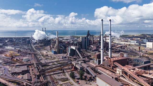 Aerial view of the Tata Steel industrial complex with tall chimneys, smoke emissions, and wind turbines along the coastline in Velsen-Noord, Noord-Holland, Netherlands.