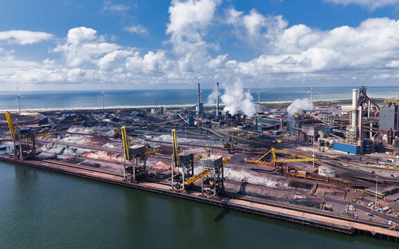Aerial view of the Tata Steel industrial complex with large cranes, raw material stockpiles, smoking chimneys, and wind turbines along the coast, Velsen-Noord, Noord-Holland, Netherlands.