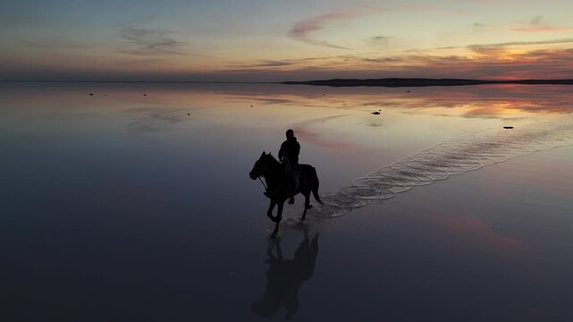Aerial view of a person riding a horse through the shallow reflective waters of Lake Tuz at sunset in Kulu, Konya, Turkiye.