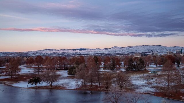 Aerial view of Ann Morrison Park with snow-covered grounds, frozen pond, and distant mountains under a soft winter sky in Boise, Idaho, United States.