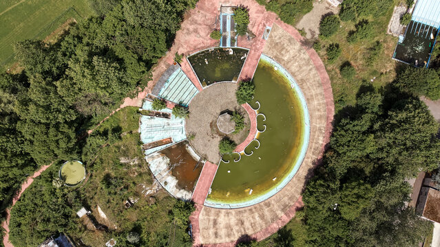 Aerial view of an abandoned circular swimming pool complex with stagnant green water, overgrown vegetation, and decaying infrastructure in Pantigliate, Lombardia, Italy.