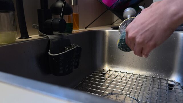 Hands washing a transparent glass coffee cup with a sponge in a modern stainless steel kitchen sink during the evening