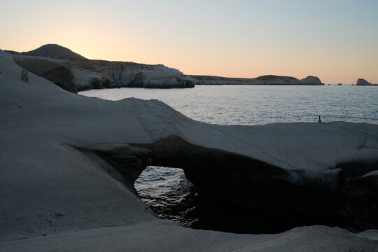 A quiet coastal landscape at sunrise, with dark volcanic rock formations silhouetted against a soft pastel sky and calm Aegean waters in Sarakiniko, Milos, Greece.
