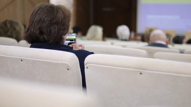 Person sits in auditorium, focused on phone. Audience members listen to presentation. Attendee captures moment on device. Useful for business, education, or tech events
