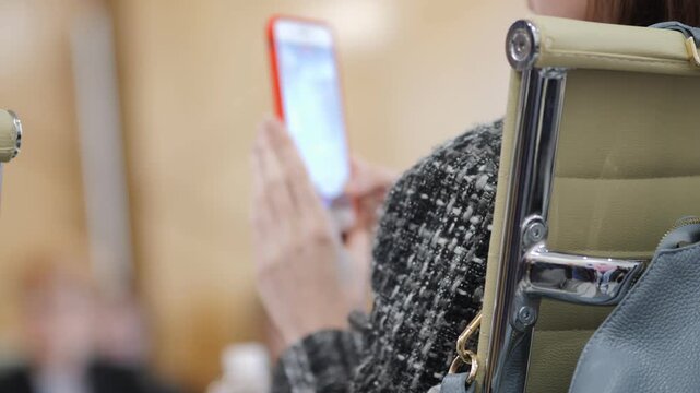 Person in chair uses phone during meeting. Focus on hands and device. Background blurred with others present. Suggests distraction or multitasking at event. Useful for themes like technology, focus