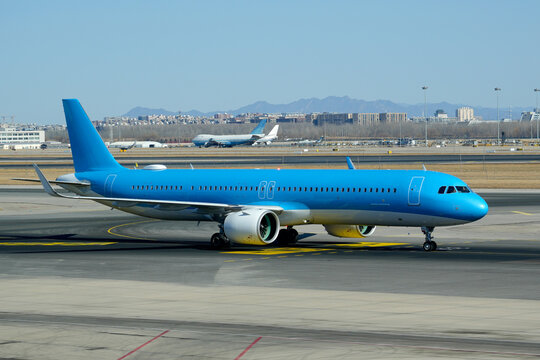 Bright blue passenger jet plane taxiing across the tarmac at a busy airport under a clear sky during the day with city buildings visible