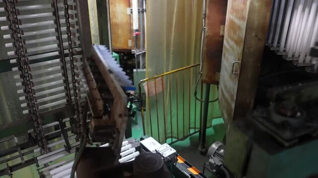 Chain conveyor carrying white coated lamp tubes past glowing machinery in a Japanese lighting factory