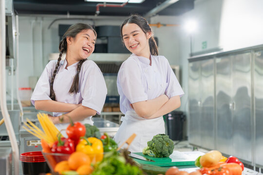 Happy young asian girl cooks smiling and laughing together in commercial kitchen, Cheerful kids in chef uniform standing with arms crossed in culinary class cooking school