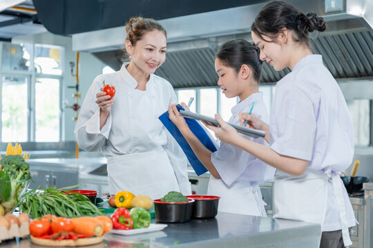 Female chef teacher explaining and teaching young students taking notes in commercial kitchen, Professional chef teaching young girls nutrition and ingredients in culinary school