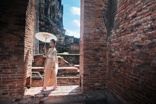 Portrait of Asian teenage girl dressed in traditional Thai outfit and umbrella with cheerful smile at old brick wall, summer holiday travel in Ayutthaya, ancient ruins heritage,  tourist destination.