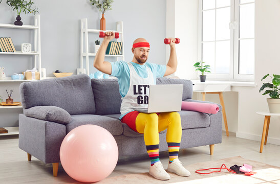 Funny man working out with dumbbells while using a laptop at home gym. Engaging in online fitness, he combines technology with exercise, adding a touch of humor to workout routine.