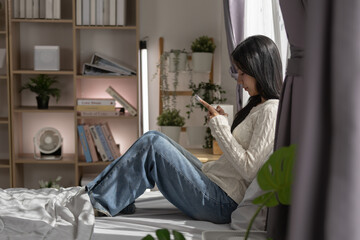 Asian woman sits on cozy bed by bright window typing message on smartphone relaxing in comfortable home bedroom feeling peaceful and focused enjoying quiet morning leisure time indoors © Ratirat