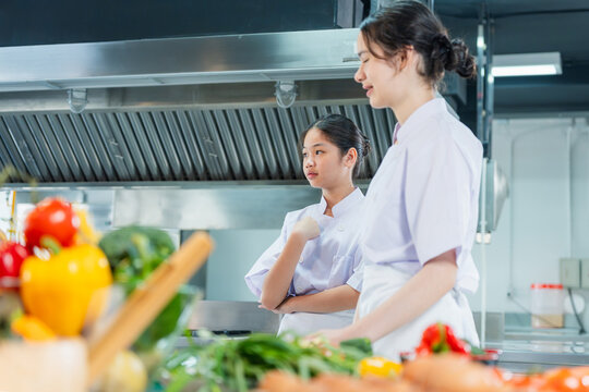 Young asian student chefs looking attentively in commercial kitchen, Teen girls in chef uniforms with fresh ingredients watching cooking demonstration in culinary school
