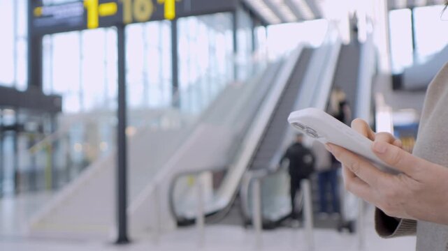 Close-up of a person holding a phone at an airport. Person focused on screen, ignoring surroundings. Escalator and terminal sign visible in background. Ideal for travel, technology, or waiting scenes